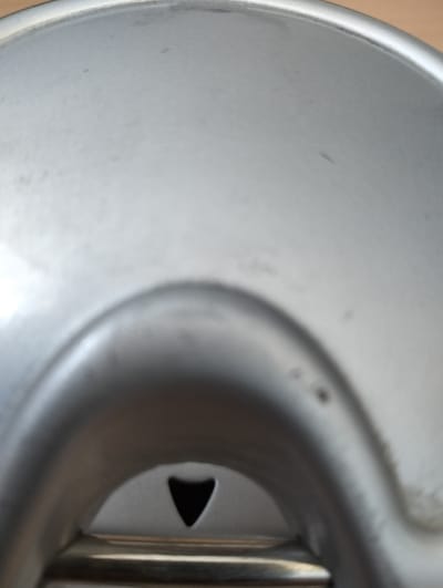 The image shows a close-up, low-angle shot of a metallic surface, possibly the lid or base of a container. The material is silver-toned and appears to be brushed metal, with subtle reflections indicating a light source from above and slightly to the left.
Dominating the lower center of the frame is a concave indentation with a dark, triangular opening or cut-out within it. This opening is deep and appears to lead into a darker space. The metallic surface surrounding this opening is curved and smooth, forming a raised lip or rim. Below this, a horizontal metallic band is visible, with a brushed texture that contrasts slightly with the main surface.
The overall environment is not discernible beyond the immediate object. The lighting is diffuse, causing soft shadows and highlights. The image does not contain any people, animals, or identifiable landmarks. The context of Qarabulaq, Kazakhstan, is not visually represented in the image itself.
