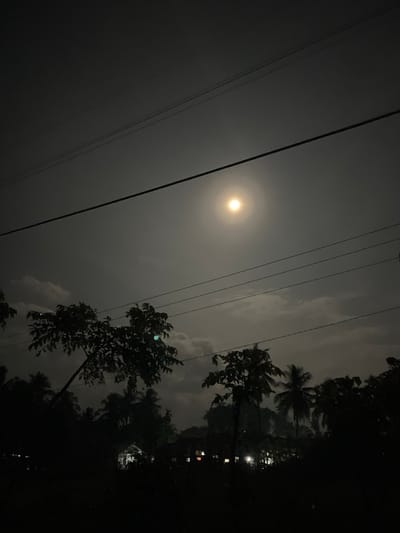 The image captures a nighttime exterior scene in Alueduamuka O, Indonesia. The sky is dark gray with prominent cloud formations partially illuminated by a bright, full moon located in the upper center-right. Multiple dark, horizontal utility lines traverse the upper half of the frame. The lower portion of the image is dominated by dense, dark silhouettes of tropical trees and foliage, including several palm trees, obscuring the ground level. In the dark background, beneath the tree line, scattered artificial lights, primarily yellowish and white, indicate the presence of residential or commercial structures. No individuals or active subjects are visible.