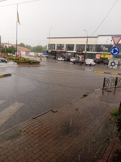 The image captures a heavy downpour in an urban setting, likely during daylight hours. The scene is dominated by a flooded intersection, with cars navigating through the water. A roundabout is visible, indicating a complex road junction. On the right side of the image, a multi-story building houses several businesses, with prominent signage for "Men Zone," "CIAO!," and "FIMKA." Other visible text includes "Brand House Kids," "Neo-mobile," and signs in Ukrainian that translate to "Fashion Shopping," "Luxury children's clothing," and "Rent and Sale of Electronics." A Ukrainian flag flies from a tall pole on the left, suggesting the location is in Ukraine. The weather is clearly rainy, with visible raindrops and puddles of water on the paved surfaces, including a cobblestone walkway in the foreground. The overall mood is somber due to the weather conditions, but the presence of businesses and traffic suggests ongoing activity.