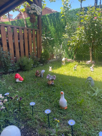 The image depicts a garden in Kraishte, Bulgaria, with a wooden fence and pergola to the left. The garden features a lawn with various decorative elements, including ceramic animal figures (goose, mouse, squirrel, rabbit), mushroom-shaped ornaments, and artificial flowers. The background includes trees and a green wall, likely a hedge or climbing plant. The scene appears to be outdoors during the day, with ample sunlight. No people are visible in the image.
