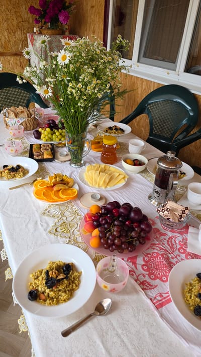 The image depicts an outdoor table setting, likely on a porch or veranda, with a spread of breakfast items. The table is covered with a white tablecloth adorned with a gold lace trim and a pink floral pattern on a portion of it.
In the center of the table, a large bouquet of white daisies and smaller white flowers in a glass vase stands prominently. To the left of the flowers, a woven basket contains sliced bread. Several small pink glass cups with floral patterns are also present.
The food items arranged on the table include plates of porridge topped with nuts and dried fruit, sliced oranges and apples, a bowl of grapes, plums, and apricots, a tray of baklava, sliced cheese, a jar of honey, a bowl of berries and fruit, and a French press filled with dark liquid, presumably coffee or tea.
Two green plastic chairs are visible on either side of the table, suggesting outdoor seating. The background shows a wooden wall and a window with a screen. The overall scene suggests a pleasant breakfast or brunch enjoyed outdoors, possibly in a rural or suburban setting given the natural elements and simple furnishings. The location is specified as Kupoyarovo, Russia. There are no people visible in the image.