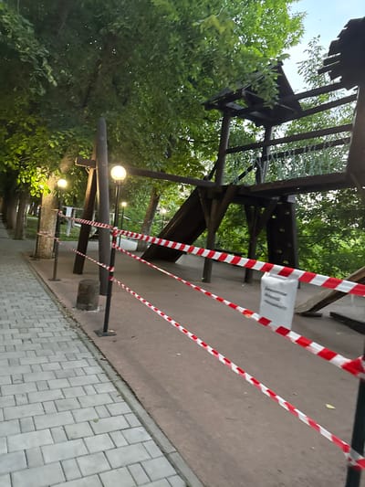 The image shows a playground or park area in Oktyabr'skoe, Russia, at dusk or early evening, as indicated by the soft lighting from several lamps. The scene is cordoned off with red and white striped caution tape, suggesting that the area is currently inaccessible or under maintenance. The tape is strung between black posts and appears to be blocking off a wooden structure, possibly a playhouse or a part of an adventure playground, which is partially obscured by lush green trees.

The wooden structure itself appears somewhat aged, with some elements looking weathered or damaged, which might be the reason for the closure. The ground in the foreground is a mix of a reddish-brown paved surface and a gray interlocking brick pathway. The caution tape creates a visual barrier, leading the eye diagonally across the frame and emphasizing the restricted access. The surrounding foliage is dense and green, indicating a pleasant outdoor environment, though the closed-off nature of the area adds a touch of somberness or disappointment.

A white sign is visible attached to the wooden structure, behind the caution tape, but the text on it is illegible. The overall impression is one of a temporarily closed recreational space, with the natural elements of trees and evening light creating a somewhat serene, yet cautionary, atmosphere.