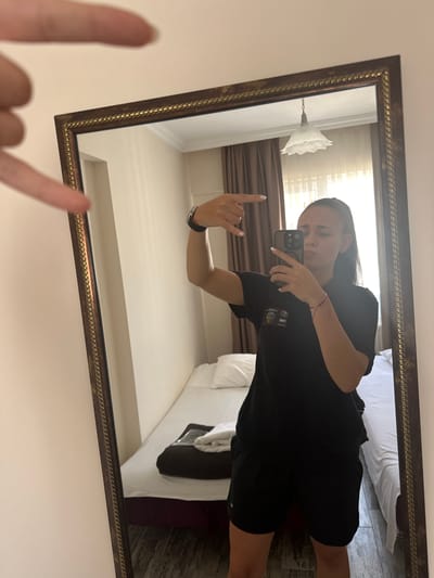 A young woman is taking a selfie in a full-length mirror. She is wearing a black t-shirt and black shorts. Her hair is dark and long, and she has a red string bracelet on her left wrist. She is holding her phone in her right hand and has her left hand raised with her index finger pointing up. The mirror is ornate with a thick, textured gold frame. The reflection shows a hotel room with a bed and curtains. The lighting suggests it is daytime. There is text on her t-shirt that is partially obscured but appears to be a logo or graphic. The location is Çanakkale Merkez, Turkey.