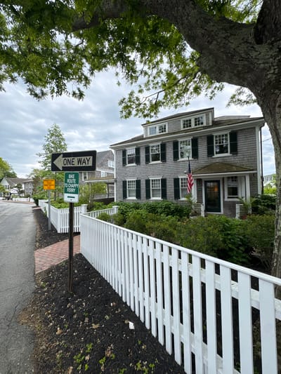 The image shows a street scene in Edgartown, Massachusetts, United States. A multi-story, grey shingled house with green shutters and an American flag displayed on its facade is centered in the background. A white picket fence runs along the right side of the image, bordering a mulched area. Street signs including "ONE WAY", "BIKE ROUTE TO CHAPPY FERRY", and a yellow traffic sign are visible along the roadside. A large tree with green foliage frames the left side of the image. The street is paved and appears slightly wet. The sky is overcast.