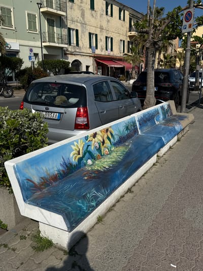 This image captures a bright, sunny street scene in Santo Stefano al Mare, Italy. The central subject is a long, ornate concrete bench painted with a vibrant mural, positioned alongside a paved sidewalk. The mural depicts an underwater world with various marine flora like corals and seaweed, transitioning to a coastal landscape with tropical-looking plants and bright yellow, large flowers that resemble sunflowers. The painting covers both the seat and the backrest of the bench.

Parked alongside the bench is a silver Kia car, its rear facing the viewer. The license plate "FB 893 YA" is clearly visible, identifying it as Italian. Items, including a cap, are visible through the car's back window. Partially obscured by the bench and a tree is a dark-colored car, possibly an SUV.

In the background, typical Mediterranean architecture is visible, with multi-story buildings featuring windows, some with closed green shutters and others with balconies. Palm trees and other green foliage line the street, casting shadows indicative of strong sunlight. A circular blue street sign with a red border and a diagonal red line, denoting a no-stopping or no-parking zone, is visible near the trees. The sidewalk is made of interlocking grey pavers, and small weeds grow along the base of the bench. The clear sky and distinct shadows suggest it is either late morning or early afternoon.