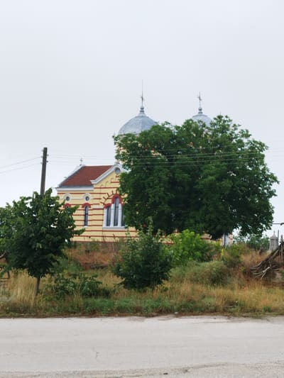 This media file displays a view of a yellow and red striped church with two silver domes against a light, overcast sky. The church features arched windows and a red roof. In the foreground, a paved road is visible, bordered by a grassy slope with scattered red flowers and several trees. A wooden utility pole with electrical wires stands to the left of the church. A large, leafy tree dominates the center and right side of the image, partially obscuring the church. The scene appears to be set in a rural or semi-rural location in Slanotran, Bulgaria, possibly during the daytime with cloudy weather. No people or specific activities are depicted, giving the image a quiet and still atmosphere. There is no visible text.
