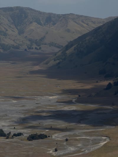 This image depicts a vast, desolate valley with a meandering riverbed, nestled between rolling hills and mountains under a cloudy sky. The landscape is characterized by dry, sparse vegetation, with patches of light-colored soil and sand creating patterns across the valley floor. The hillsides are covered in a mix of scrub and scattered trees, with some areas appearing more densely forested. Shadows from the clouds cast dramatic contrasts across the terrain, highlighting the contours of the land. The overall impression is one of a remote, rugged, and somewhat barren natural environment. This scene is consistent with the landscape found in the vicinity of Cemorolawang, Indonesia, particularly the caldera of Mount Bromo, known for its "Sea of Sand" and surrounding volcanic hills.