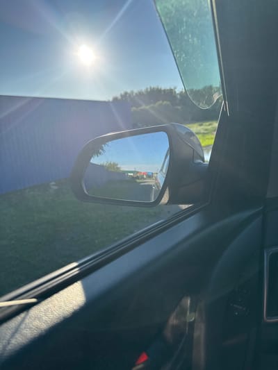 The image is taken from inside a vehicle, looking out the passenger side window and at the side mirror. The sun is shining brightly in the upper left corner of the frame, casting a lens flare. In the distance, visible through the side mirror, is a road with several colorful objects, possibly vehicles or containers, lined up. The side mirror reflects a clear blue sky and some trees. Outside the car, a blue wall or fence runs along the left side of the frame, with green grass in front of it. The interior of the car is visible in the foreground, with the black door panel and a hint of the window controls. The image appears to be taken in a rural or semi-urban environment on a sunny day. The specific location in Starokhalilovo, Russia, is not evident from the visual information provided in the image.