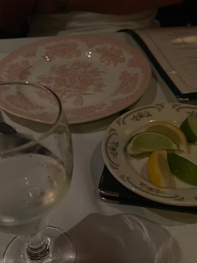 The image displays a close-up of a dining table setting, presumably within a restaurant or formal establishment in University Park, United States. A white tablecloth covers the table surface. In the foreground, a clear, stemmed glass containing a transparent liquid is partially visible on the left side, exhibiting reflections. Adjacent to it, a small, white, round plate with a subtle dark floral border pattern holds two yellow lemon wedges and two green lime wedges. This plate rests on a dark, rectangular object. Further back and centrally located is a larger, empty, round white plate featuring a prominent pink floral toile pattern. To the right of the large plate, a dark-bound menu or booklet is partially visible. The upper portion of the background is out of focus, revealing indistinct shapes, including a person wearing a light-colored top behind the large plate. The scene is illuminated by ambient light, casting reflections on the glassware and plates. No active interactions are depicted.