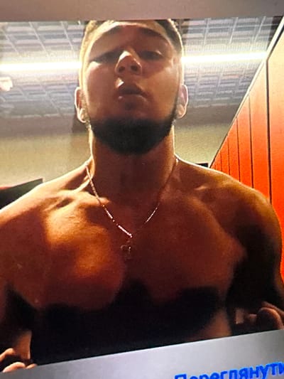 The image shows a close-up of a shirtless man with a beard, wearing a necklace. He is standing in front of a row of bright orange lockers. The background also shows a ceiling with lights. The image appears to be taken in a gym or locker room. There is no indication of the city of Siedlce, Poland, within the image itself or its context.