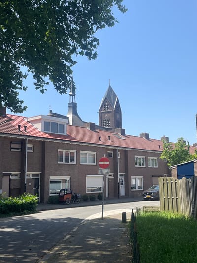 This image depicts a street scene in Venlo, Netherlands, on a clear, sunny day. The perspective is from a higher vantage point, looking down and slightly to the right.
The primary subjects are a row of brick apartment buildings with red-tiled roofs. One building features a prominent clock tower with a dark spire, possibly part of a church. In front of the buildings, a street runs parallel to them. Parked on the side of the street is a dark-colored car. There is also a three-wheeled cargo bike with a brown cargo box.
Green trees and lush grass are visible, indicating a pleasant, possibly summer, season. The sky is a bright, cloudless blue. A traffic sign with a red circle and a horizontal bar, indicating "no entry" or a similar restriction, is visible on a pole along the street. A smaller sign below it shows a bicycle icon with the text "uitgezonderd" beneath it, suggesting an exception for cyclists.
The scene appears to be a residential area, with the architecture and street layout being characteristic of Dutch towns. The lighting suggests it is daytime, likely late morning or early afternoon, given the strong sunlight and shadows cast. There are no people actively engaged in activities, giving the impression of a quiet moment in the day. The overall mood is serene and ordinary, capturing a typical street view.