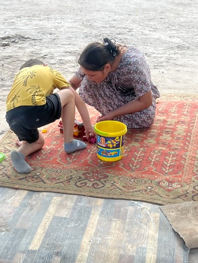 In the city of Balkh, Tajikistan, a young child wearing a yellow t-shirt with a palm tree print and black shorts, and light gray socks, kneels on a rug. Beside them, an adult woman in a floral dress is also kneeling. They are both engaged with a yellow toy bucket and building blocks. The rug is red with a patterned design and is placed on a surface of wooden planks. The background appears to be a dirt or gravel ground.