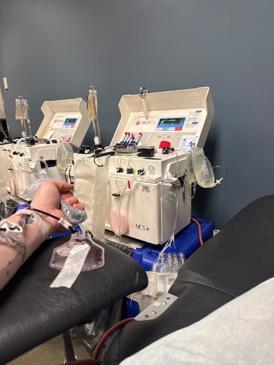 A person undergoing apheresis sits in a dark chair. A bag of blood is taped to the armrest. The arm has tattoos and an IV. A large white machine is attached to the IV line. There is a clear bag of liquid hanging near the machine. A blue container sits behind the machine. The wall is gray. There are two identical machines next to each other.