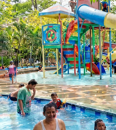 The image depicts a water park scene in Durin Simbelang, Indonesia. In the foreground, several people are standing in the water. One adult is smiling at the camera while others, including children, are partially submerged and playing. In the background, a colorful water play structure features slides and platforms. Trees surround the water park, contributing to a lush, tropical environment. Other individuals are visible in the background, both in and out of the water.