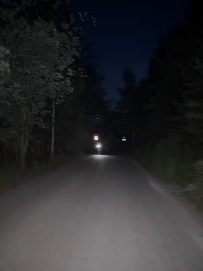 The image depicts a dirt road at night in a forested area, possibly near Dom Otdykha Zolotoy Plyazh, Russia. The road is dark and appears to be unpaved, with trees lining both sides. In the distance, a vehicle's headlights are visible, illuminating the road ahead. There are also some lights and structures on the right side of the road in the distance, which are unclear due to the low light conditions. The sky is dark, indicating it is nighttime.