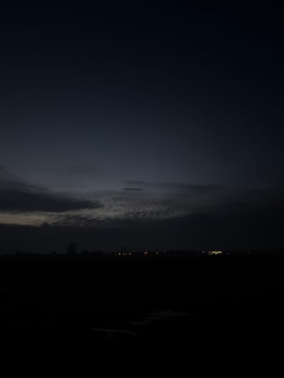 A dark, nighttime image reveals a cityscape's faint lights along the right edge. Ominous dark clouds dominate the left, with streaks of faint, lighter clouds. The overall tone is dark and atmospheric, suggesting either a stormy night or a low-light environment where the city's illumination is minimal.