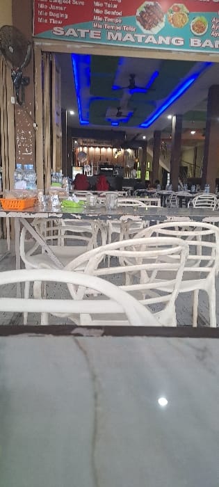 The image depicts the interior of a casual dining establishment, likely a restaurant, located in Alue Mudem, Indonesia. The perspective is from a table in the foreground, looking inward.
In the immediate foreground, a blurred, reflective light-colored surface, possibly a marble or polished concrete tabletop, is visible. The midground features multiple white, intricately designed plastic chairs, some partially obscured and appearing empty. A long, weathered white table with a grey top and visible peeling paint is prominent. On this table, multiple clear drinking glasses, several small clear plastic bottles of water, an orange plastic basket containing unidentified items, and a green object (possibly a small container or napkin holder) are arranged.
The background shows the interior of the restaurant. The ceiling features recessed sections with bright blue LED strip lighting and green panels, along with at least two ceiling fans. A large red banner above the entrance displays menu items, including "SATE MATANG BANG" in large white letters, and lists various "Mie" (noodle) dishes such as "Mie Telur," "Mie Seafood," "Mie Tempe," and "Mie Tempe Telor," accompanied by images of food. To the left, a dark-colored wall-mounted fan is visible. Further inside, multiple tables and chairs are arranged, most appearing unoccupied. At least two individuals are visible in the deeper background; one figure is seated with a red garment partially visible, while another figure is less distinct. A dark, tall appliance, possibly a refrigerator or counter, is also present within the restaurant's interior. The overall lighting is artificial and bright.