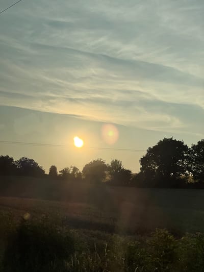 The image depicts a rural landscape at sunset. The sky is filled with wispy clouds, illuminated by the setting sun, which appears as a bright orb with a lens flare. The sun is positioned slightly to the left of the center, casting a warm, golden light across the scene. In the foreground, silhouetted trees form a dark line against the bright sky. Beyond the trees, a field stretches out, its details obscured by the low light conditions. Power lines are visible running horizontally across the middle ground, a subtle indication of human presence in the otherwise natural setting. The overall impression is one of tranquility and the fading light of day. The location is stated to be the city of Derry/Londonderry, United Kingdom, suggesting this is a view from the outskirts of the city, looking out into the countryside.