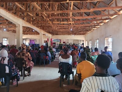 An indoor scene at a large gathering in Rinze, Nigeria, depicts numerous individuals seated and facing a stage area. The room features light-colored, unadorned concrete walls with multiple rectangular windows fitted with metal security grates. The ceiling exposes a wooden truss roof structure with visible rafters, beams, and what appears to be corrugated metal sheeting directly above. Several white-painted concrete support columns punctuate the space. The floor is an unpaved or concrete surface.
Approximately 50 to 70 people, primarily adults and some children, occupy the majority of the seating area, arranged in rows on individual plastic chairs. Most individuals are dressed in varied traditional African patterned garments, with many women wearing head coverings. They are oriented towards the front of the room, suggesting an audience for an event.
At the far end, a slightly raised stage is present. A purple fabric serves as a backdrop. On the stage, at least one large screen displays illuminated text or graphics. A person is visible standing behind a podium or desk. To the right of the stage, another smaller screen or monitor is positioned. Sound equipment, including speakers, is visible on the stage. To the left of the stage, an individual is operating what appears to be sound mixing equipment. Overhead lighting fixtures are sparsely distributed within the exposed roof structure.