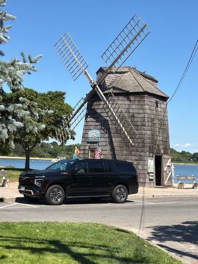 A black Chevrolet Suburban is parked on a paved road in front of a weathered wooden windmill. The windmill has two large, open wooden blades and a shingled roof. A sign on the windmill reads "Village of Sag Harbor Information Center." An American flag and a colorful flag are mounted on the windmill. A body of water, likely a bay or harbor, is visible behind the windmill, with trees and distant hills in the background. The sky is clear and blue. A patch of green grass is in the foreground, with shadows from trees cast upon it. There is a white bench to the left of the windmill. The license plate of the vehicle is partially visible and appears to be from New York.