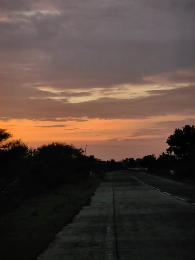The image shows a rural road in Dattapur, India, at dusk. The sky is filled with dramatic clouds painted in shades of orange and purple, indicating the setting sun. The road, paved with asphalt and a concrete median, stretches into the distance, bordered by dark silhouettes of trees and bushes on either side. A few white markers or small walls are visible along the side of the road. The lighting is dim, suggesting either early morning or late evening. There are no people or vehicles visible in the scene.