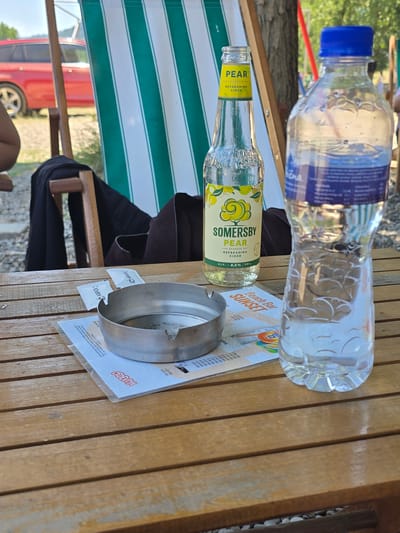 A medium shot shows a wooden table at an outdoor venue in Zvornik, Bosnia and Herzegovina. On the table are a bottle of Somesrby Pear refreshing cider, a plastic bottle of water with a blue cap, an ashtray, and a menu. In the background, a red car is parked near a green and white striped deck chair. The ground is covered with gravel.