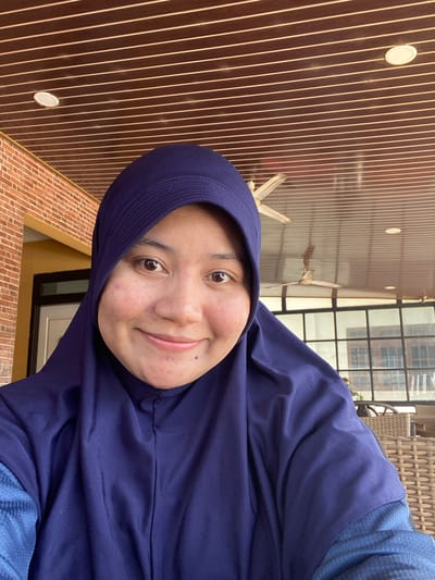 In an outdoor selfie, a young Indonesian woman wearing a navy blue hijab smiles warmly at the camera. She has a visible mole on her chin and some minor blemishes on her cheeks. Her eyes are dark brown and expressive. The background of the image suggests a covered outdoor patio or veranda, possibly part of a residence in Keraton, Indonesia. The ceiling is made of wooden slats with white trim, and a ceiling fan is visible overhead. To the left, a brick wall with a yellow accent can be seen. Through a large window behind her, a glimpse of patterned glass and woven furniture indicates a comfortable seating area. The lighting suggests it is daytime, with the sun illuminating the scene.