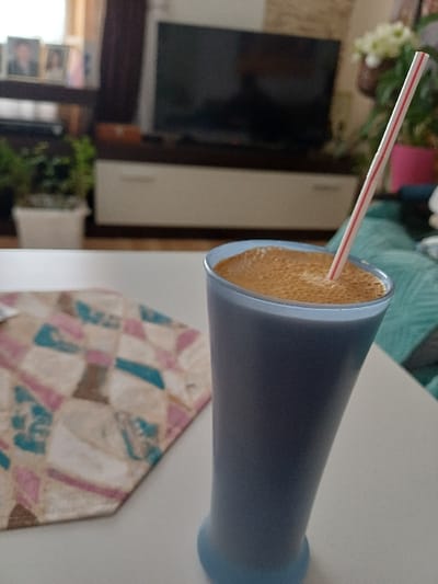 A light blue-tinted glass, containing a dark beverage topped with a layer of light brown foam, stands on a white surface in the foreground. A red and white striped straw is inserted into the drink. To the left of the glass, a multi-colored fabric placemat with a geometric pattern (pink, blue, brown, beige) is partially visible. The background is a blurred indoor setting, likely a living room. A large, dark-screened television is mounted on a white and brown console unit. To the left of the television, a window is visible, and several framed photographs are displayed on a shelf or windowsill. Green potted plants are also present in the background, one in a white pot on the left, and another with white flowers in a pink pot on the right. No individuals are visible. This scene is located in a residential interior in the city of Prdejci, North Macedonia.