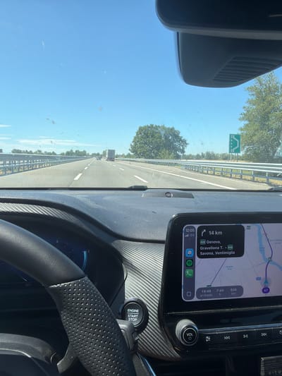 This is a first-person view from inside a car traveling on a highway in Italy, likely on a sunny day. The dashboard is visible, featuring a touchscreen display showing a navigation app with a route to Ventimiglia. The app indicates 14 km remaining to the destination, with an estimated arrival time of 19:49 and a travel time of 7 hours and 44 minutes, covering a total distance of 836 km. Other elements on the dashboard include a volume knob, media control buttons, and an engine start/stop button. The steering wheel, wrapped in perforated leather, is also visible. Outside the car, the highway stretches ahead, bordered by a metal guardrail and trees. A road sign with an icon of a bridge is visible on the left side of the highway. The sky is clear and blue, suggesting good weather. The time displayed on the navigation screen is 12:05, and the 5G signal is active. The text "Castelnuovo Scrivia" and "Torrente Grue" are visible on the map, confirming the location context.