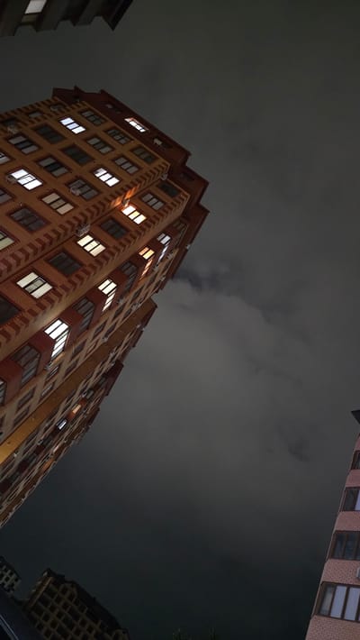 This image depicts a nighttime view of apartment buildings in Khasavyurt, Russia, looking upwards towards a dark, cloudy sky. The primary subjects are the multistory residential buildings, characterized by their brick facades and numerous illuminated windows. The architecture suggests a dense urban environment.

The scene captures the quiet atmosphere of a night in a city. No people are visible, and the focus is on the structures themselves and the sky above. The activity is the passive observation of urban life at night, with the lights in the windows indicating ongoing habitation within the buildings.

Notable details include the varying degrees of light emanating from the windows, some glowing brightly, others more subtly. The clouds in the sky are heavy and dark, suggesting overcast weather or potentially the early stages of rain, contributing to the dimness of the scene. The angle of the shot, from a low perspective looking up, emphasizes the scale and height of the buildings. The overall mood is somewhat somber and quiet, typical of a dark, cloudy night. There is no visible text in the image.