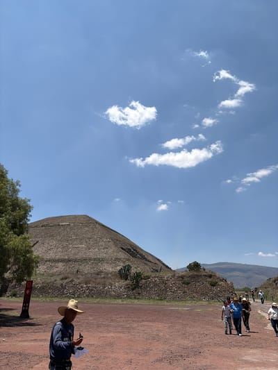 The image shows an outdoor scene in San Sebastián Xolalpa, Mexico, with the Pyramid of the Sun prominently featured. The pyramid is a large stone structure, partially obscured by vegetation. A group of tourists is present on a dirt path beside the pyramid. A person wearing a straw hat and dark clothing is standing on the right side of the frame, appearing to be either reading from papers or taking notes. The sky above is blue with scattered white clouds.