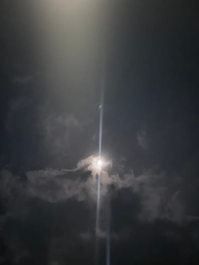 This image, taken in Lehvaz, Armenia, depicts a nocturnal sky. A bright celestial body, identifiable as the moon, is partially obscured by dark, patchy cloud formations, primarily in the lower-middle portion of the frame. The moon emits a strong light, generating a distinct vertical lens flare that bisects the image. A subtle green artifact is discernible within the upper segment of the flare. The scene is devoid of human subjects, man-made objects, or discernible terrestrial features, focusing exclusively on atmospheric and celestial elements under ambient dark conditions.