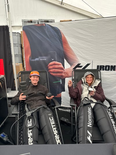 Within a white event tent in Weymouth, United Kingdom, two individuals, a male and a female, are seated in black mesh reclining chairs, facing the camera and smiling. Both subjects have their legs encased in black "Normatec" compression boots, which are actively in use.
The male, positioned on the left, is wearing a dark grey zip-up jacket and an orange, white, and blue baseball cap. He holds a black foam roller in his left hand and a small black massage ball in his right hand.
The female, on the right, wears a maroon padded jacket over a light grey hooded top with a black and white striped hood lining. She holds a white and black coffee cup in her left hand and raises her right hand with an open palm gesture.
Behind them, a large promotional banner displays an arm holding a "Hypervolt GO" massage gun and part of the "IRONMAN" logo. Stacks of grey plastic storage crates are visible in the background, behind and to the left of the banner. The tent's internal framework is visible overhead.