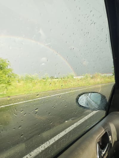 From inside a moving vehicle, a view through a rain-streaked window reveals a segment of a rainbow in the sky. The road visible is wet and marked with white lines. A side-view mirror of the car is also visible. Outside the window, blurred green foliage lines the roadside. Based on the city name, it is presumed to be in Bogdanovo, Bulgaria, though specific location details are not discernible from the image.