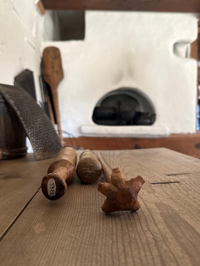The image depicts a close-up view of wooden objects on a rustic wooden table in what appears to be a historical setting. In the foreground, three wooden tools are arranged on the table. The closest object on the left is a cylindrical tool with a lighter-colored wooden handle and a darker tip, bearing a white label with the number "3326". To its right is another cylindrical wooden object, possibly a pestle, with a cracked surface. Further to the right is a small, intricately carved wooden object resembling a star or a flower with multiple points.
In the background, a traditional stove, likely a Russian stove (pech), is visible. The stove is white-washed and has a dark arched opening where cooking pots are placed. To the left of the stove, a grater and another wooden utensil, possibly a spatula, are leaning against the wall. The environment suggests a historical dwelling or museum exhibit, potentially related to traditional Russian life and cuisine, likely located in Shushenskoye, Russia, a region known for its historical significance, particularly in connection with Vladimir Lenin's exile. The lighting is soft, and the focus is on the foreground objects, with the background slightly blurred, creating a sense of depth.