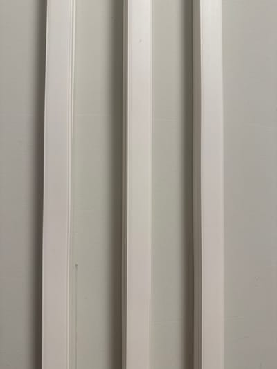 The image displays a close-up, eye-level, full-frame view of three vertical, white decorative moldings against a light gray, textured wall. The moldings are evenly spaced and appear to be made of wood or a similar material. The lighting creates subtle shadows, highlighting the relief and texture of the moldings and the wall. No people, specific city locations, or actions are depicted. The image focuses solely on the architectural details.
