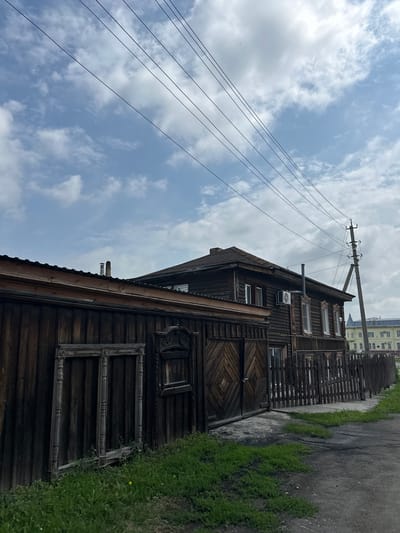 The image depicts a rustic wooden house with a fence in Nogornyi, Russia. The house is made of dark-stained wood, featuring multiple windows with white frames and an air conditioning unit. A prominent wooden gate with a diamond pattern is visible, as well as a detached wooden window frame leaning against the fence. The fence itself is made of vertical wooden planks. In the background, a utility pole with wires and a distant yellow building can be seen. The ground is a mix of dirt and patchy green grass. The sky is partly cloudy with a pale blue hue. No people or actions are apparent in the scene.