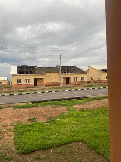 The image depicts a partially constructed or damaged building with a yellow facade and orange trim, situated next to a road. The building's roof appears to be under construction or has been damaged, with exposed metal beams visible. Several windows with white frames are present on the facade. The road is paved and has a black and white striped curb. To the right of the road is a grassy area with some patches of bare earth. The sky is overcast with clouds. The scene is captured from an elevated viewpoint, looking down at the building and road. The overall impression is one of ongoing construction or recent damage. There is no visible text in the image. The location is specified as Dumnga, Nigeria. The time of day appears to be daytime, given the ambient light, and the weather seems to be cloudy.