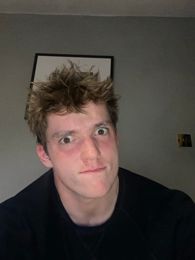 A young Caucasian male with short, tousled brown hair, wearing a black crew neck sweater, looks directly at the camera with a somewhat confused expression. His face is illuminated by a light source to the left of the frame, casting shadows on the right side of his face and hair. There is a framed picture on the wall above him, with the wall painted a light grey color. The image appears to have been taken indoors, likely in a bedroom or living room, with a wall switch visible in the background. The location of this image cannot be determined from the coordinates provided, as they correspond to a general area in Telford, rather than a specific address or building.