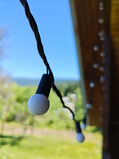 The image shows a string of outdoor lights against a bright blue sky and a backdrop of green trees and foliage. The string of lights is black and twisted, with individual white bulbs attached. One bulb in the foreground is in sharp focus, showing a textured, speckled surface and a black cap. Other bulbs further down the string are out of focus, appearing as blurred white circles.
To the right of the frame, a wooden structure, possibly part of a porch or roof, is visible. The light suggests it is daytime, and the clear sky indicates fair weather. The scene evokes a sense of leisure and outdoor enjoyment, potentially a backyard or patio setting. The location is identified as Ukeshi, Georgia. No people or text are visible in the image.