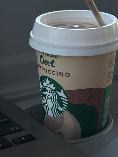 A close-up photograph shows a Starbucks Oat Cappuccino in a white-lidded cup with a straw, resting on a laptop keyboard. The Starbucks logo and parts of the "Oat Cappuccino" text are visible. The cup's design includes green and brown patterns. The location is not identifiable as Reggio nell'Emilia, Italy, from the image content.