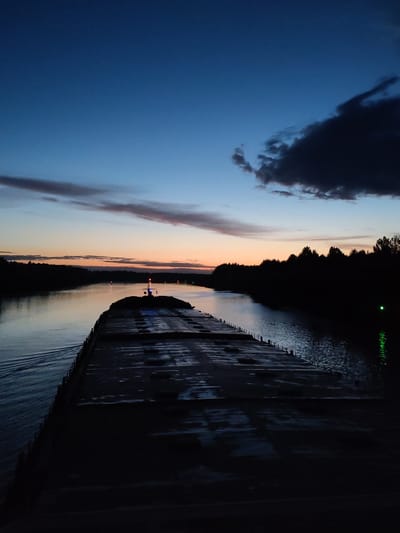 The image depicts a barge moving on a river at dusk. The sky is a gradient of blue fading to orange near the horizon. There are clouds present in the sky, some are dark and prominent. The river reflects the colors of the sky. On either side of the river are dark silhouettes of trees. The foreground is dominated by the dark, wet surface of the barge. A small structure is located towards the front of the barge. No people are visible. The location is not possible to determine solely based on the visual elements of the image and cannot be verifiably determined from the city of Uzhla, Russia.