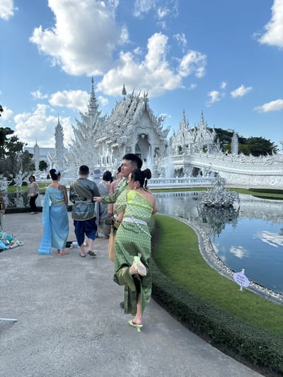 The image depicts a daytime scene at Wat Rong Khun, commonly known as the White Temple, in Ban Rong Khun, Thailand. The temple, an elaborately ornate structure rendered entirely in white with intricate carvings and spires, dominates the background under a bright blue sky dotted with scattered white cumulus clouds. A reflective pond or moat with an ornate white sculpture in its center runs along the right side of the temple, reflecting the sky and clouds. Manicured green grass and low hedges line the edge of the pond.
In the foreground and midground, a group of individuals, primarily wearing traditional Thai attire, walks along a paved concrete path. A woman in a vibrant green one-shoulder top and a matching wrap-around skirt with a diamond pattern, adorned with a white belt and light green high-heeled mules, is captured in motion, turning her head towards the left while her right leg is lifted behind her, showing the sole of her shoe. Beside her, a man in a green patterned traditional shirt and dark blue pants walks, extending his left arm slightly forward. Further left, a woman in a light blue traditional one-shoulder outfit with a gold belt and flat sandals walks away from the camera, revealing a tattoo on her left shoulder blade. Other individuals in similar attire are visible behind them, along with a woman in casual attire on the far left. On the far left ground, a light blue object, possibly fabric or a bag, is visible, along with a partial white structure. A white sign with Thai script and "NO ENTRY" in English is placed at the edge of the lawn near the pond on the right.