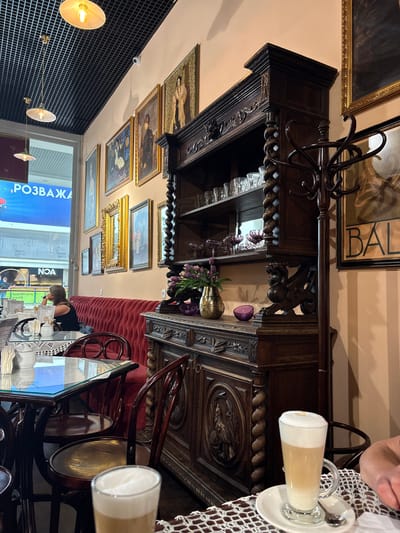 The image depicts the interior of a cafe, likely located in Sokilnyky, Ukraine, given the presence of Cyrillic text on a nearby sign reading "РОЗВАЖА". The cafe has an old-world ambiance with ornate dark wood furniture, including a large display cabinet filled with glasses and decorative items. The walls are adorned with several framed paintings, suggesting an artistic theme. Seating includes a plush red velvet couch and dark wooden chairs with curved backs. A woman is seated on the couch, looking towards a window or display area. On a table in the foreground, a glass of latte with foam art is visible, along with a white ceramic sugar dispenser and a white lacework placemat. The lighting appears to be warm, with decorative pendant lamps hanging from a black grid ceiling.