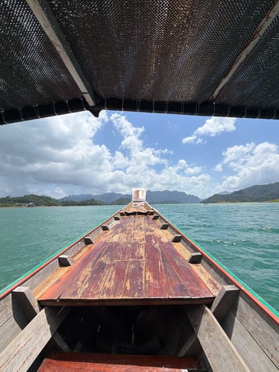 The image depicts a point-of-view shot from the front of a wooden longtail boat traveling on a body of water, likely in Ban Chiao Lan, Thailand. The boat has a worn, reddish-brown wooden deck with visible grain and some damage. The sides of the boat are also made of wood, with a green stripe along the top edge. Above the passengers, a dark mesh canopy is stretched over a frame, providing shade.

The water is a vibrant turquoise or teal color and appears slightly choppy, with small waves reflecting the sunlight. In the distance, a lush, green landscape of forested hills and mountains rises from the water. The sky is partly cloudy, with white cumulus clouds against a clear blue sky. The overall impression is a serene and scenic journey through a tropical environment. No people are visible in the frame.