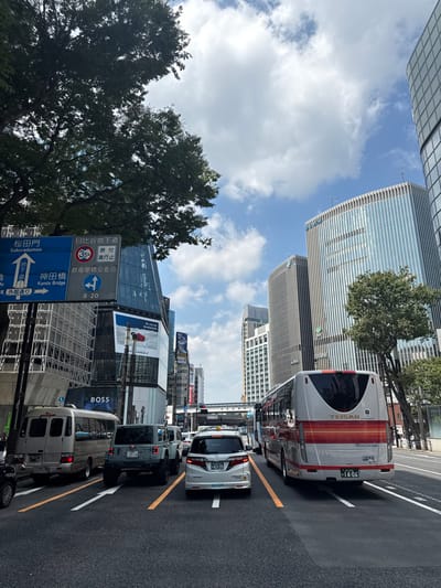The image depicts an urban street scene on Sotobori-dori, within Chūō Ward, Tokyo, Japan, observed looking west towards the adjacent Otemachi area of Chiyoda Ward. The scene is illuminated by daylight under a clear, partially cloudy sky.

The multi-lane asphalt road shows moderate traffic, with vehicles in all three visible lanes appearing stationary or moving slowly. The lanes are demarcated by white dashed lines, with a solid yellow line defining the leftmost edge. In the leftmost lane, a light-colored commercial van is followed by a grey two-door Jeep Wrangler (license plate: 足立 580 ほ 25-35). The middle lane is occupied by a silver Honda Odyssey minivan (license plate: 品川 502 せ 43-55). The rightmost lane contains the rear of a white and orange "TEISAN" branded coach bus (license plate: 品川 200 か 14-06).

On the left side of the street, a large tree with dense green foliage partially obscures the upper sky and background buildings. Below it, a blue overhead street sign with white arrows and text provides directions: "桜田門 Sakuradamon" (straight arrow), "神田橋 Kanda Bridge" (right arrow), "日比谷地下道 Hibiya Underpass" (left/down arrow, indicating 35 km/h limit and "原付通行止" - No Moped Traffic), and "大手町交差点 Otemachi Intersection" (right arrow). Adjacent to the sign, a modern angular glass building features a large billboard displaying an Air France advertisement and a prominent "BOSS" retail sign at street level.

On the right side of the street, multiple high-rise buildings dominate the skyline. The most prominent is a tall structure with a facade of vertical blue-tinted glass, displaying the "SMBC" (Sumitomo Mitsui Banking Corporation) logo. Behind it, another tall building with a grey, vertically-lined facade is visible. A smaller tree with green foliage is present at street level on the right. An elevated road or railway structure is visible in the distance over the street. No individuals are distinctly visible or identifiable.