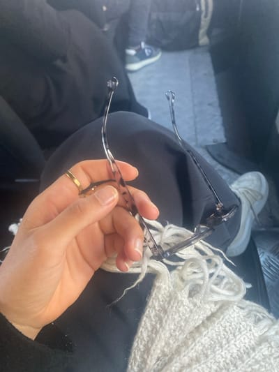 The image displays an interior scene, likely within a vehicle, focused on a person's left hand holding a pair of folded eyeglasses. The hand is light-skinned, with a plain gold-colored band worn on the fourth digit. The fingernails are short and unpolished. The eyeglasses have a dark tortoiseshell pattern on their frames and temples, with transparent, rounded tips on the earpieces. The temples are extended upwards and backward from the main frame. Below the hand, a light-colored, possibly off-white, knitted or crocheted textile with fringed edges is draped over the person's lap, which is covered by dark fabric, likely trousers. In the right mid-ground, a blurred portion of a second individual's right foot is visible, wearing a white sneaker with a thick sole and prominent laces. This foot rests on a textured, possibly ribbed, metal floor panel. Further into the background, indistinct dark clothing of another seated person is discernible. The specific location within Monastir, Tunisia, is not ascertainable from the visual information provided in the image.