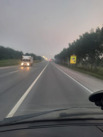 The image captures a view from inside a vehicle driving on a highway. The road is wet, indicating recent rain or damp conditions, and the sky is overcast and hazy, suggesting it might be dusk or dawn. A white truck with its headlights on is visible in the distance, traveling in the same direction as the camera vehicle. On the right side of the highway, there is a line of trees and a yellow rectangular sign with some symbols on it, possibly indicating a speed limit or other traffic information. The foreground shows the dashboard and windshield of the vehicle, with some marks and a crack visible on the windshield. The general atmosphere is muted and slightly foggy. The location is specified as the highway on the outskirts of Berd', Russia.