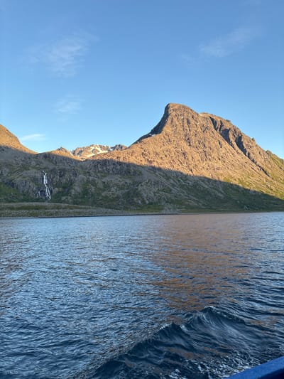 This is a photograph of a coastal mountain landscape taken from a boat. The foreground consists of dark blue water with small waves. In the midground, a rocky, mountainous shoreline is visible, with a waterfall cascading down the side of a hill on the left. The mountains are partially illuminated by sunlight, casting shadows. The peaks in the distance are snow-capped. The sky is clear and blue with a few wispy clouds. The scene is likely captured during a sunset or sunrise, given the lighting. The location is Arnøyhamn, Norway.