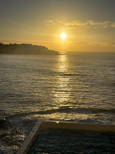 The image depicts a sunrise over the ocean in Bondi Beach, Australia. The sun, low on the horizon, casts a golden light across the water, creating a shimmering effect. The coastline is visible, with buildings silhouetted against the sky. In the foreground, a swimming pool is partially visible, with waves crashing against its edge. The image captures a tranquil coastal scene at dawn.